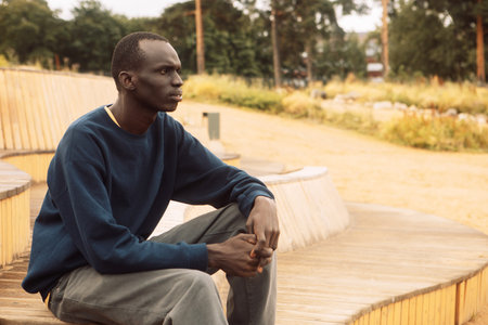 A dark-skinned young man sits on a wooden bench in the park and listens to music. Lifestyle and people concept.の写真素材