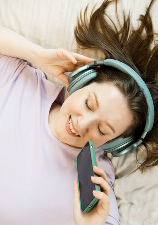 Pretty adult woman lies on the bed in headphones and listens to music with her eyes closed and holding a smartphone in her handsの写真素材