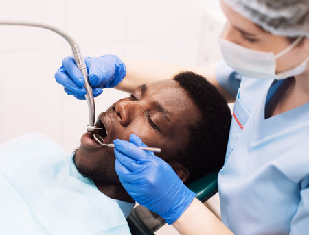 Portrait of black male patient getting teeth treatment with dentist at modern clinic, sitting in chair in white cabinet. professional stomatologist female using dental drill tool for young man.の写真素材