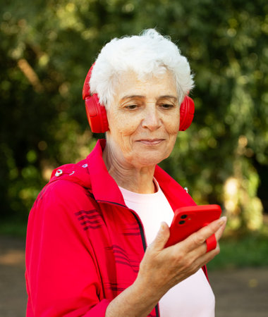 An elderly woman with a short haircut dressed in red sportswear listens to music on headphones, using a smartphone while jogging. summertime.の写真素材