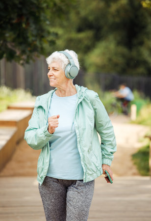 An elderly woman with a short haircut dressed in sportswear is jogging in the park while listening...