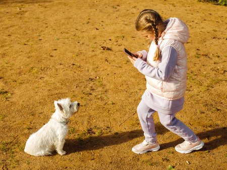 A seven-year-old girl with pigtails plays with a white dog on the beach and takes pictures of her...