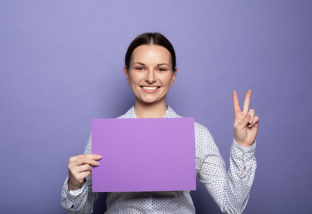 Young brunette woman holding purple blank advertising board standing on lilac background in studioの写真素材