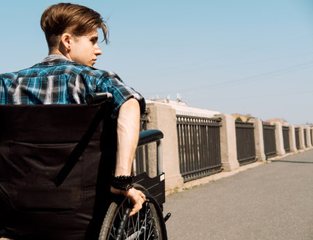 A young disabled man rides in a wheelchair across a bridge, the young male dressed in a plaid shirt and jeans. view from the back.の写真素材