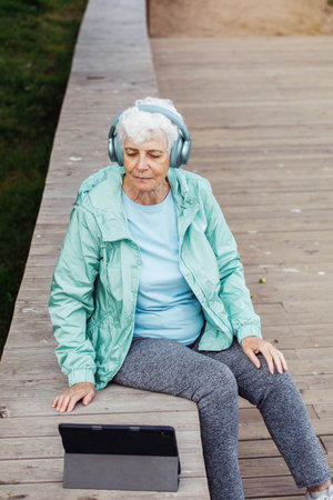 An elderly woman with short hair sits on a park bench and uses a tablet. The pensioner listens to music on headphones and uses the Internet.の写真素材
