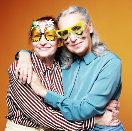 Two elderly women friends wearing party glasses hugging on yellow background. Lifestyle, friendship...
