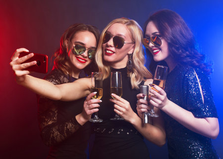 Group of partying female friends clinking flutes with sparkling wine and holding microphone and mobile.の写真素材