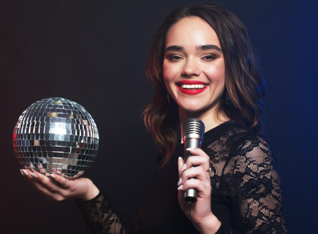 young woman wearing black dress, holding disco ball and singing into microphone over dark backgroundの写真素材