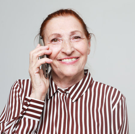 Old smiling woman wearing striped shirt talking on cell phone over gray background. Emotion, technology and old people concept.の写真素材