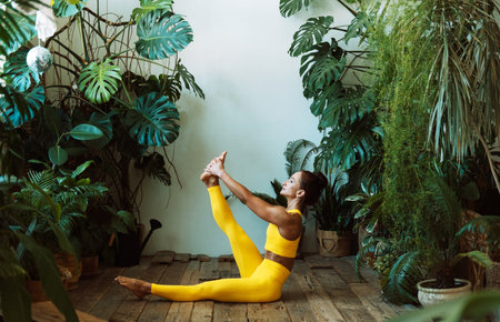 Brunette woman practices yoga at home among flowers. Urban jungle and gymnastics, yoga, pilates concept.の写真素材