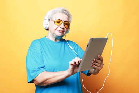 Education, old people and technology concept: Elderly woman in headphones with tablet over yellow...