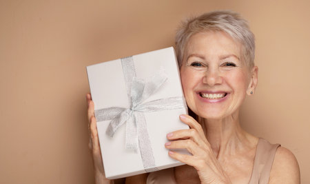 Photo of smiling lady pensioner holding present box isolated beige color backgroundの写真素材