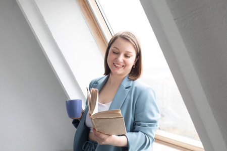 Smiling adult woman reading book and drinking coffee near window at home