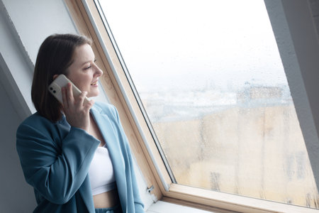 Happy adult woman talking on mobile phone near window. Lifestyle and technology concept.の写真素材