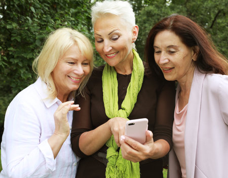 Three old women are smiling and looking photos at the at screen of the phone in the park on a summer dayの写真素材