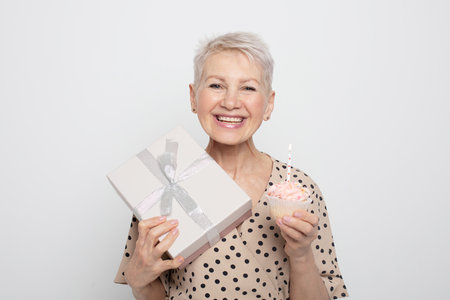 charming pensioner woman with short haircut holding a gift and a cupcake with a candle, celebrating a birthdayの写真素材