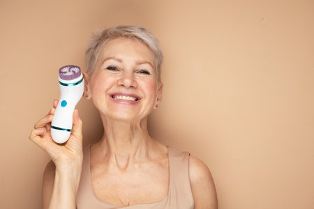 Beautiful 60s woman holding a massager in her hand over beige background. Spa technique skincare ads.の写真素材