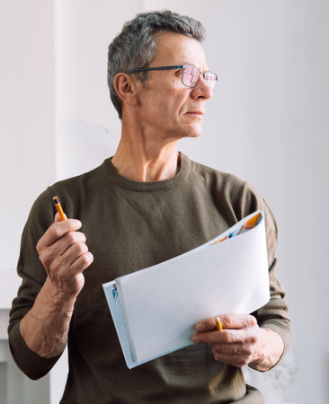 A man holds an album and a pencil, makes sketches. The man is gray-haired and dressed casually,...