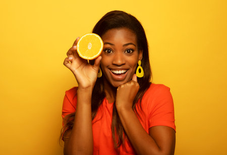 Healthy summer, diet and people concept: Cheerful afro woman holding orange over yellow background.の写真素材