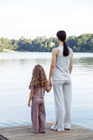 A young mother and her five-year-old daughter watch the fish in the lake, standing on the bridge. Back view. Family and people concept.の写真素材