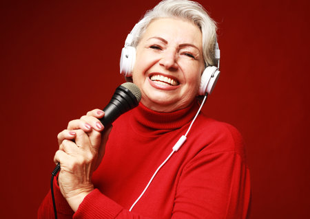 Happy senior woman singing with microphone, having fun, expressing musical talent over red backgroundの写真素材