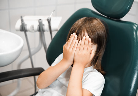 Pleasant little girl lying in a dentist chair and covering her eyes, being afraid of the checkup at the dentistsの写真素材