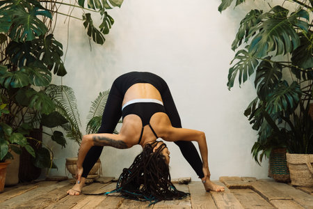Young woman with braids practices yoga at home among flowers. Urban jungle and gymnastics, yoga,...