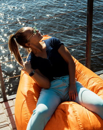 A stylish young woman is relaxing while sitting in a bean bag chair on the embankment near the...