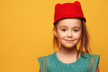 Stylish little girl model in a red hat and blue dress on a yellow background. Fashion and people concept.の写真素材