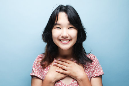 Portrait of sincere brunette Asian female model smiles broadly expresses happiness feels joyful has white even teeth wears pink dress isolated over blue backgroundの写真素材
