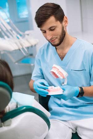 Young Male dentist shows dentures with a bracket in hands to the patient. False teeth in the hands