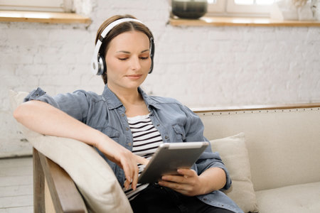 Relaxed young woman in casual clothes and headphones watching movie on tablet while sitting on sofa...