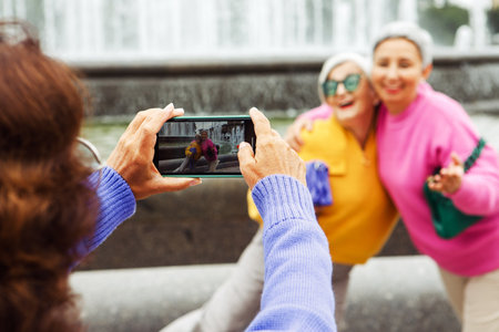 Three cheerful elderly friends in bright sweaters take a selfie on a smartphone and laugh, having a great time. Happy pensioners. Summer in the city.の写真素材