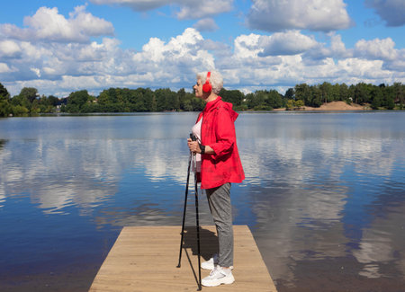 An elderly woman with short hair, dressed in a red jacket, stands with Nordic walking poles on a bridge by the lake nd listening music with headphones.の写真素材