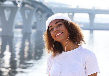 Young cute afro american woman wearing white t-shirt listening to music in headphones near sea. Summer time.の写真素材