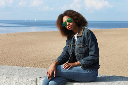 Beautiful african woman in a denim jacket and sunglasses is resting while sitting on the beachの写真素材