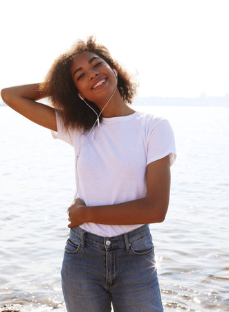 Young cute afro american woman wearing white t-shirt listening to music in headphones near sea. Summer time.の写真素材