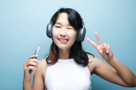 Cheerful smiling Asian teenage girl listens to music on headphones and shows a victory sign over blue background.の写真素材