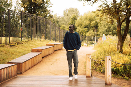 Portrait of a young handsome African man dressed in a blue sweatshirt, holding a backpack, walking in the park on a summer dayの写真素材
