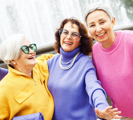 Three happy cheerful pensioner female friends in bright sweaters and sunglasses walk together....