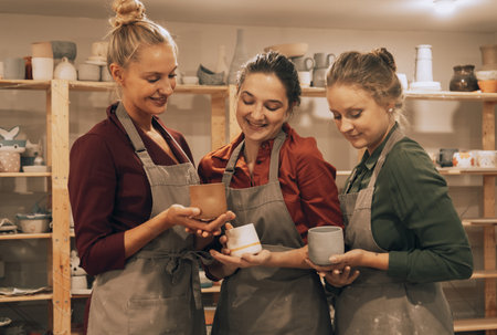 A company of three female friends in a ceramic workshop shows each other homemade mugs. The concept...