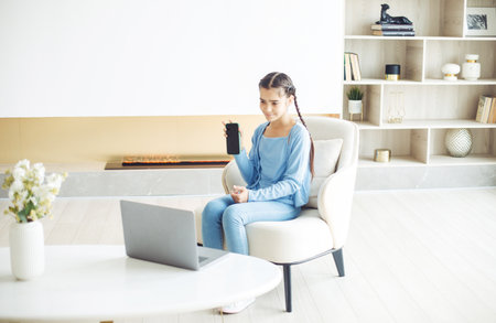 A schoolgirl with pigtails sits in a chair in a modern living room and works with a laptop, lifestyle conceptの写真素材