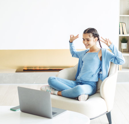 A schoolgirl with pigtails sits in a chair in a modern living room and works with a laptop, lifestyle conceptの写真素材