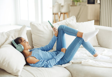 Little kid cute girl sitting on sofa, using smartphone with headphones, take selfie. Kids and gadgets concept.の写真素材