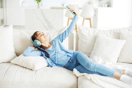 Little kid cute girl sitting on sofa, using smartphone with headphones, take selfie. Kids and gadgets concept.の写真素材
