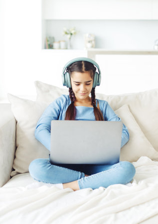 Schoolgirl with pigtails wearing headphones sits on the sofa and works with a laptopの写真素材