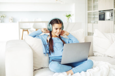 Schoolgirl with pigtails wearing headphones sits on the sofa and works with a laptopの写真素材