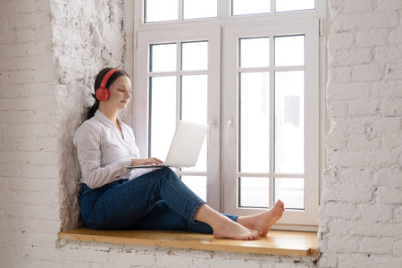Young woman sitting at the window at home with laptop and red headphones