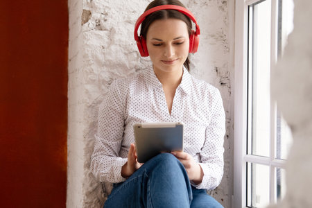 Young woman sitting at the window at home with digital tablet and red headphones