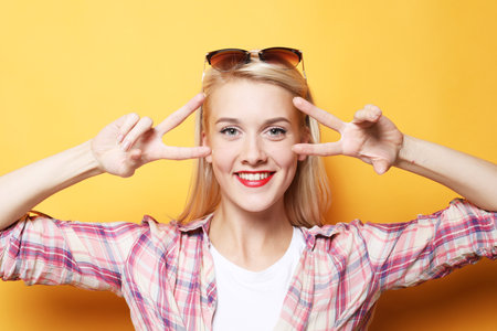 Portrait of a happy young blond woman showing victory gesture and winking, studio shoot over yellow backgroundの写真素材